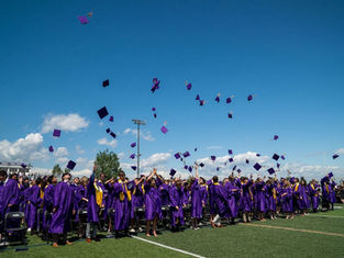 Graduates in purple gowns toss caps into a bright blue sky on a football field, celebrating at an outdoor ceremony.
