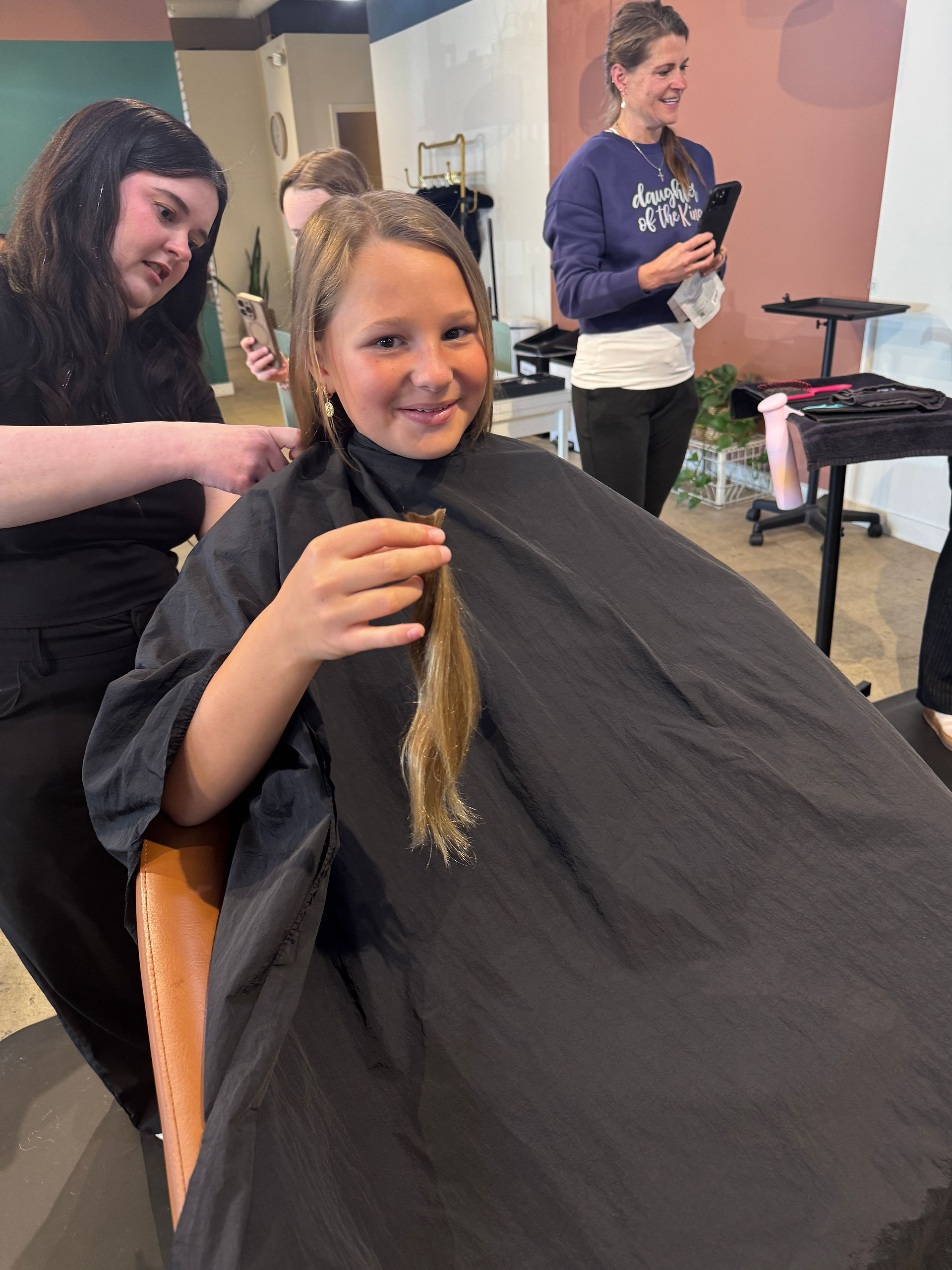 Girl in a salon chair holds a cut section of her hair while a stylist trims the rest.