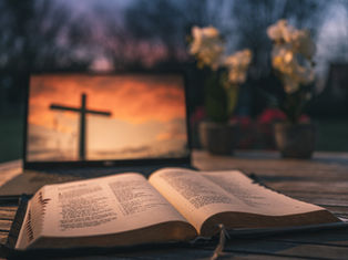 Open Bible on a wooden table in front of a laptop showing a cross at sunset, with blurred flowers and a warm, reverent mood