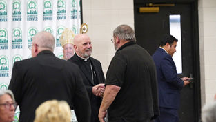 Cleric in black robe shakes hands with a man in a black shirt. Archdiocese of Denver banner in background. Another man uses phone.