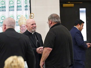Cleric in black robe shakes hands with a man in a black shirt. Archdiocese of Denver banner in background. Another man uses phone.