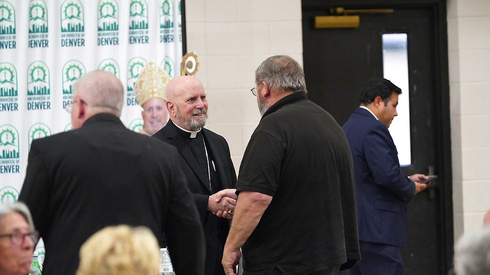 Cleric in black robe shakes hands with a man in a black shirt. Archdiocese of Denver banner in background. Another man uses phone.