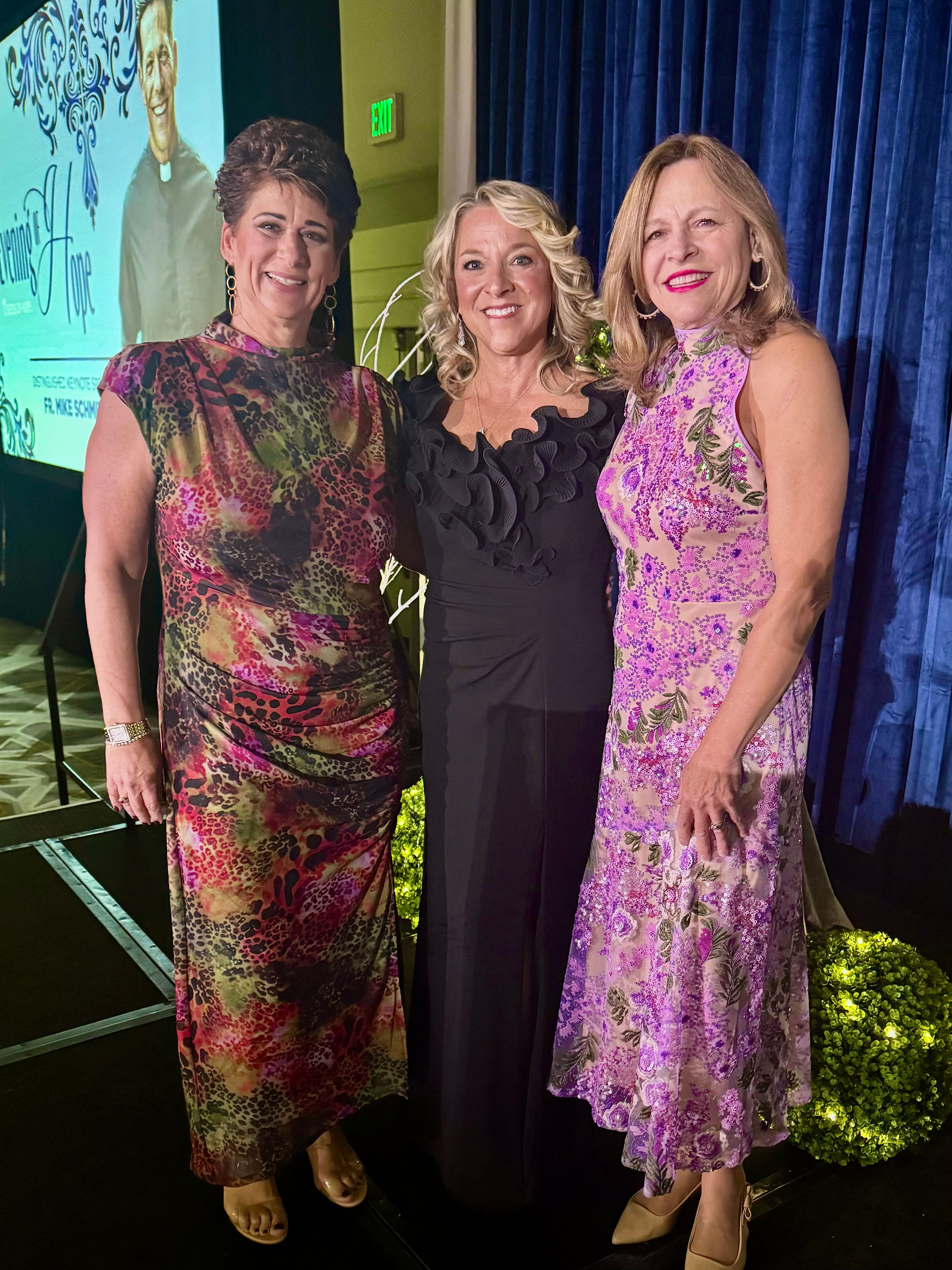 Three women in elegant dresses smile in front of a blue curtain at an event. A poster in the background reads "Evening of Hope."