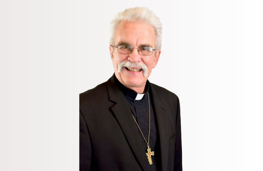 Elderly man with glasses and mustache smiling, wearing a black suit and clerical collar with a cross necklace, on a plain white background.
