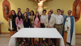 A group of people smiling in front of an altar with a crucifix. Candles are lit. The cloth reads, "This is my body, this is my blood."