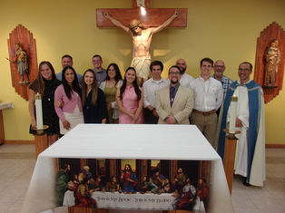 A group of people smiling in front of an altar with a crucifix. Candles are lit. The cloth reads, "This is my body, this is my blood."