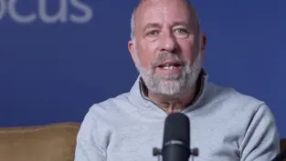 Man with a beard speaking into a microphone against a blue background with partial "CUS" text visible. He wears a gray sweater, seated on a brown couch.