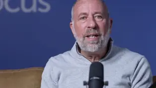 Man with a beard speaking into a microphone against a blue background with partial "CUS" text visible. He wears a gray sweater, seated on a brown couch.