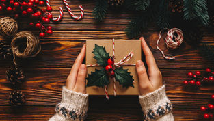 Hands in cozy sweaters hold a gift wrapped with holly and candy cane twine on a wooden table surrounded by festive decor and pinecones.