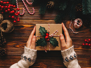 Hands in cozy sweaters hold a gift wrapped with holly and candy cane twine on a wooden table surrounded by festive decor and pinecones.