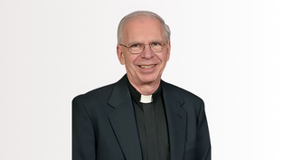 Smiling man in clerical attire with a white collar and glasses, set against a plain white background.