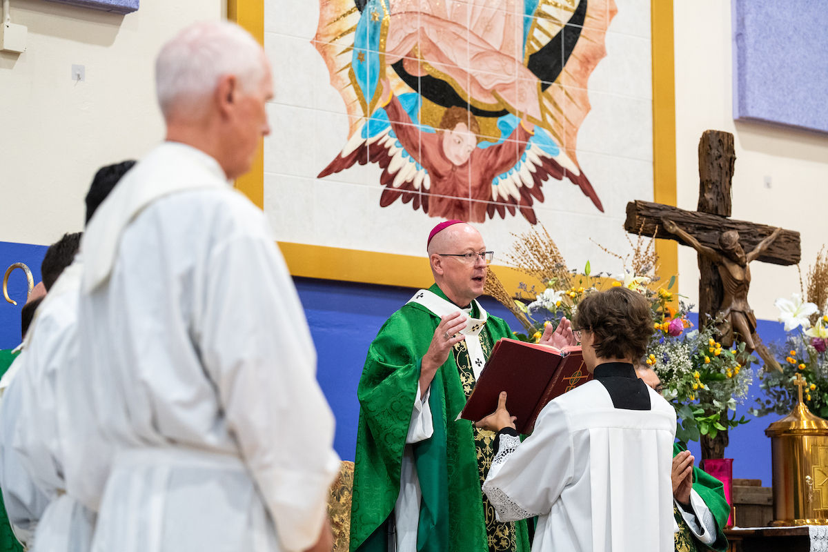 Catholic bishop in green vestments blesses a book during Mass, with altar servers, crucifix, and flowers in a church.