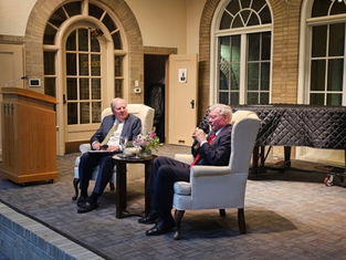 Two men in suits sit in armchairs on stage, engaged in a discussion. A podium and covered piano are in the background. Cozy indoor setting.
