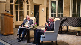 Two men in suits sit in armchairs on stage, engaged in a discussion. A podium and covered piano are in the background. Cozy indoor setting.