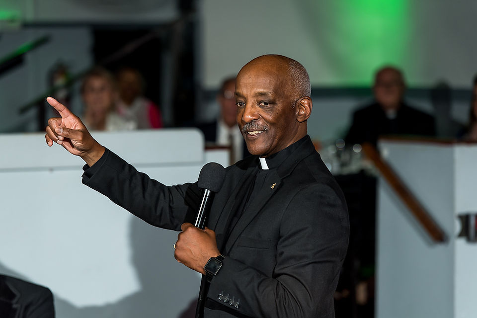 Smiling man in a black clerical outfit holding a microphone and pointing. Blurred audience in the background with green lighting.