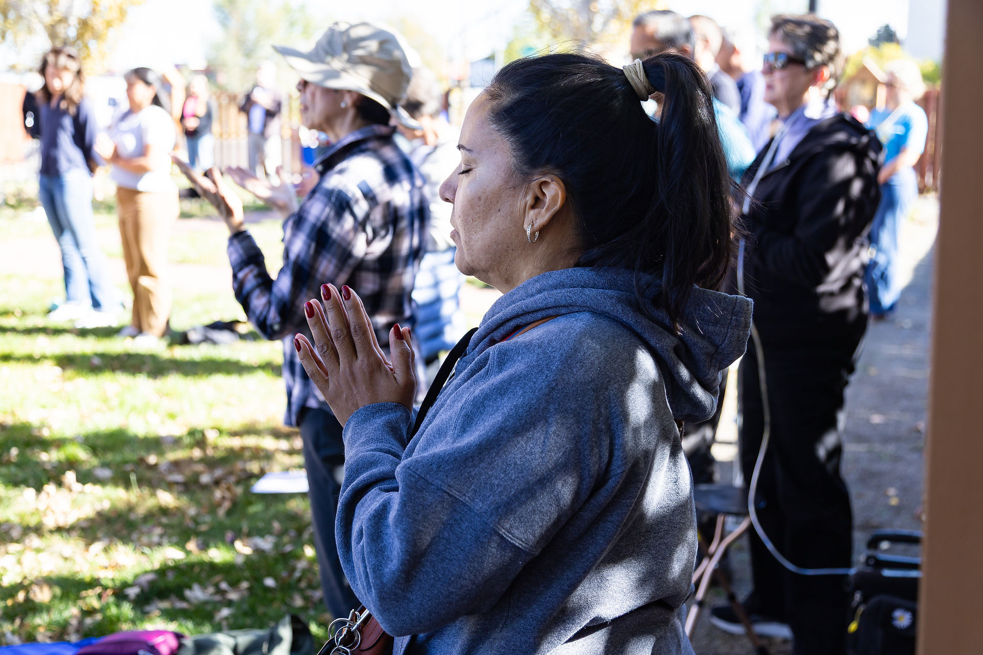 Woman in gray hoodie prays outdoors with eyes closed, surrounded by people standing on grass. Sunlit scene with trees in background.