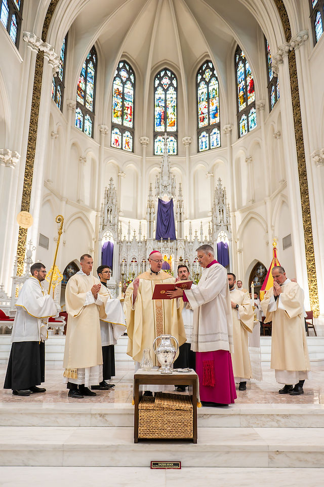 Clergy in white robes conduct a ceremony in a grand cathedral with stained glass windows. A book is held open, creating a solemn atmosphere.