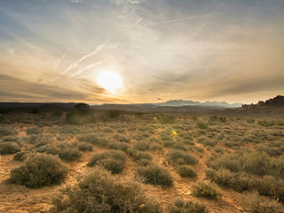 Sunset over a desert landscape with sparse bushes, distant mountains, and a vapor trail in the sky, creating a warm, tranquil scene.