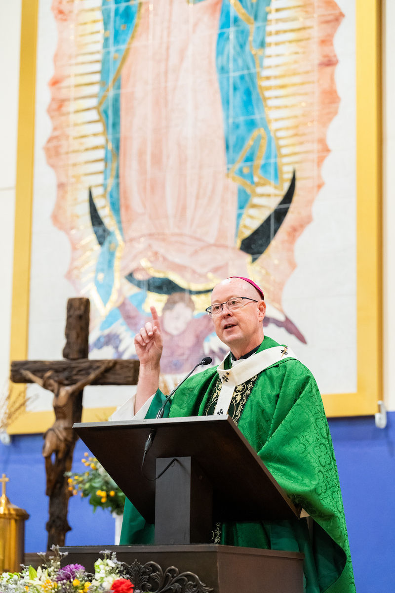 Bishop in green vestments speaks at a church lectern, raising one finger, with a crucifix and large Virgin Mary image behind.