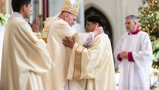 A bishop in ornate robes hugs a priest warmly in a church. Two other clergymen watch. Background shows soft lights, creating a serene mood.
