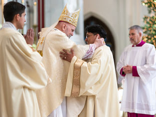 A bishop in ornate robes hugs a priest warmly in a church. Two other clergymen watch. Background shows soft lights, creating a serene mood.
