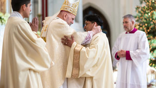 A bishop in ornate robes hugs a priest warmly in a church. Two other clergymen watch. Background shows soft lights, creating a serene mood.