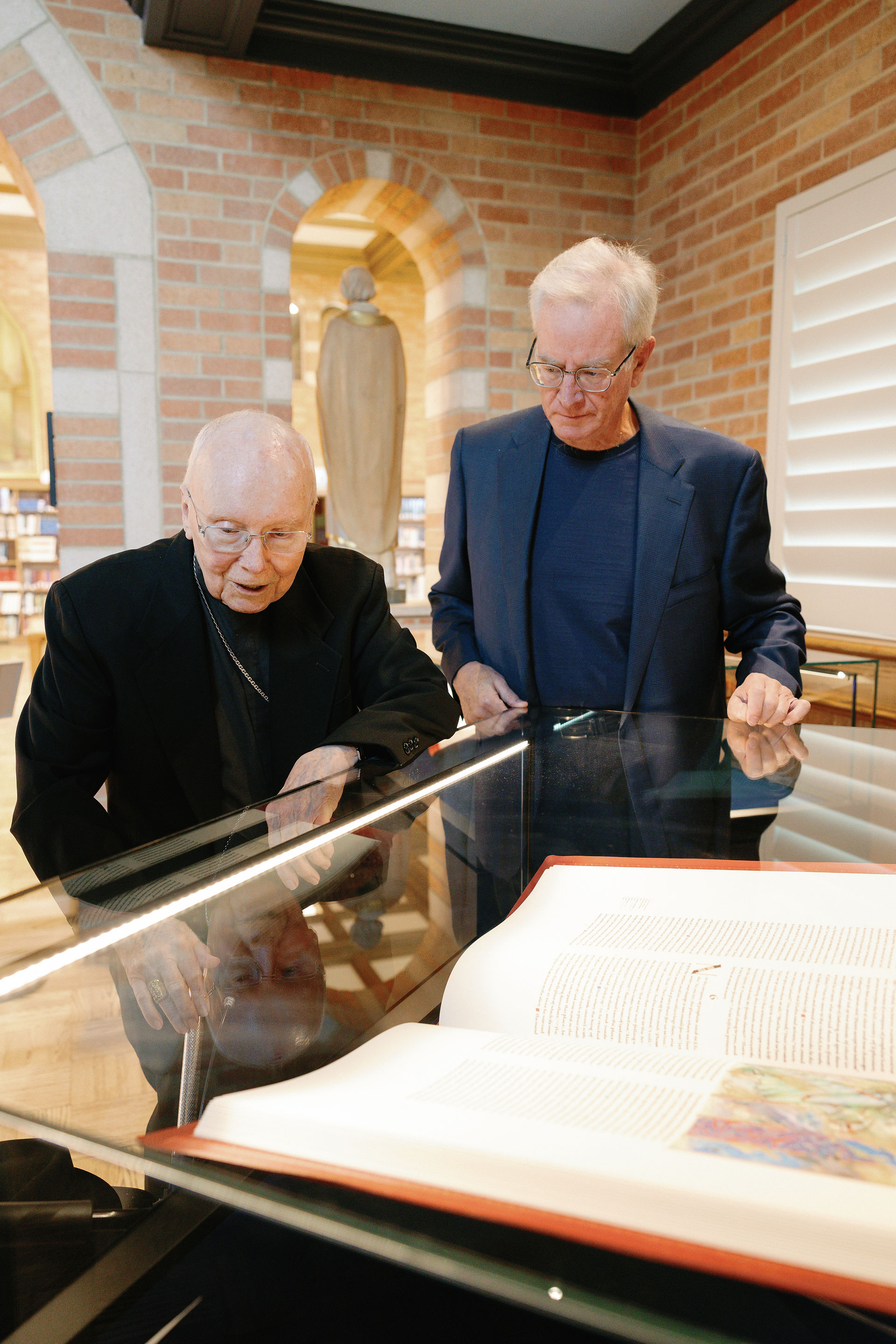 Two men in a library examine a large, open book inside a glass case. The brick walls and archways create a historic ambiance.