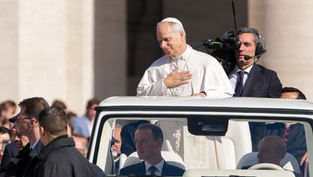 A man in white robes, standing in a vehicle, touches his chest, surrounded by people. A cameraman films nearby. Bright, outdoor setting.