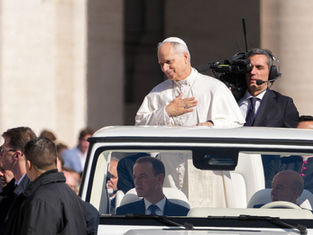 A man in white robes, standing in a vehicle, touches his chest, surrounded by people. A cameraman films nearby. Bright, outdoor setting.