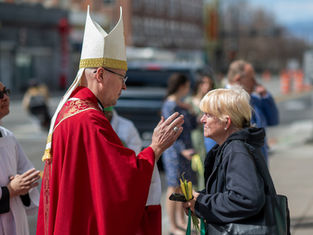 A man in a red robe and white mitre is blessing a woman holding palm fronds on a city street, surrounded by onlookers.