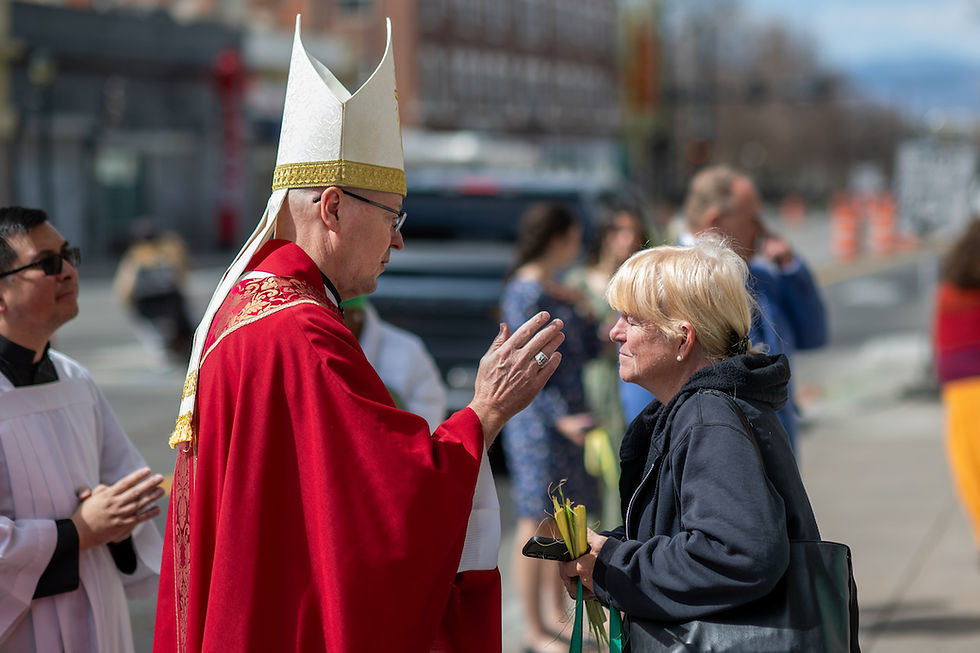 A man in a red robe and white mitre is blessing a woman holding palm fronds on a city street, surrounded by onlookers.