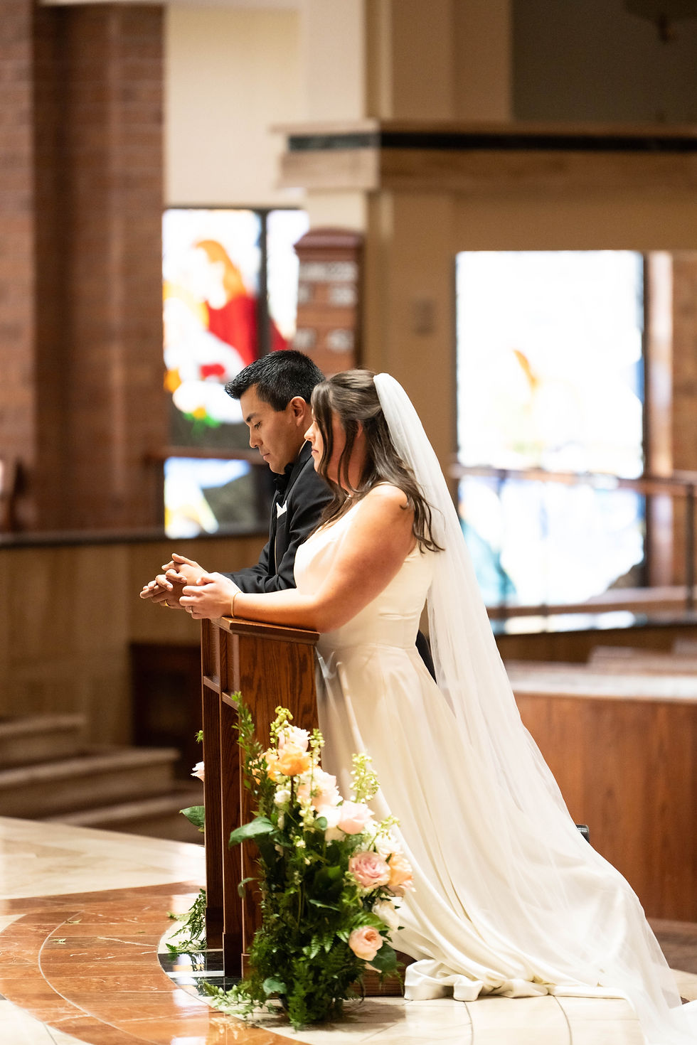Bride and groom pray at a wooden altar adorned with flowers in a church. The scene is serene, with colorful stained glass in the background.