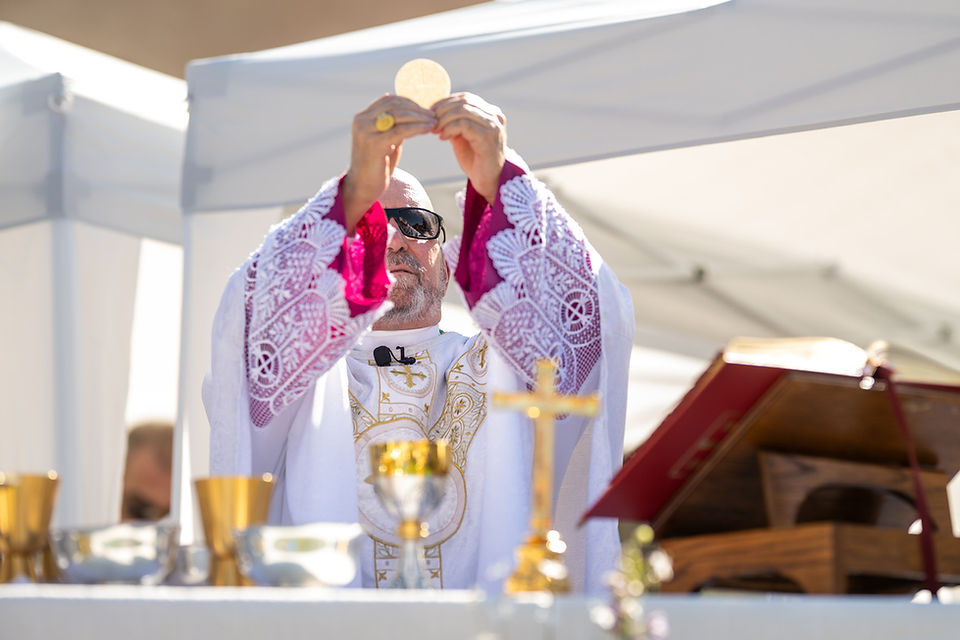 Priest in white and magenta vestments lifts a communion host at an outdoor Mass under a white canopy, with chalice and cross on altar.