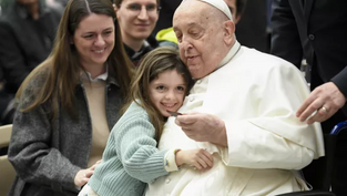 Child in a green sweater hugs an elderly man in white attire, seated in a wheelchair. Others around smile warmly. Indoor setting, joyful mood.
