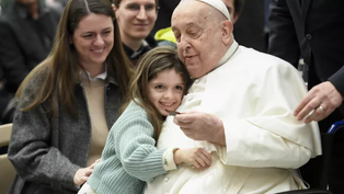 Child in a green sweater hugs an elderly man in white attire, seated in a wheelchair. Others around smile warmly. Indoor setting, joyful mood.