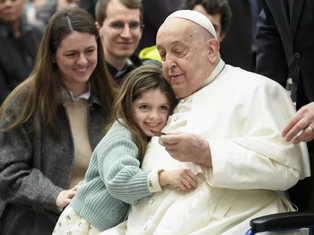 Child in a green sweater hugs an elderly man in white attire, seated in a wheelchair. Others around smile warmly. Indoor setting, joyful mood.