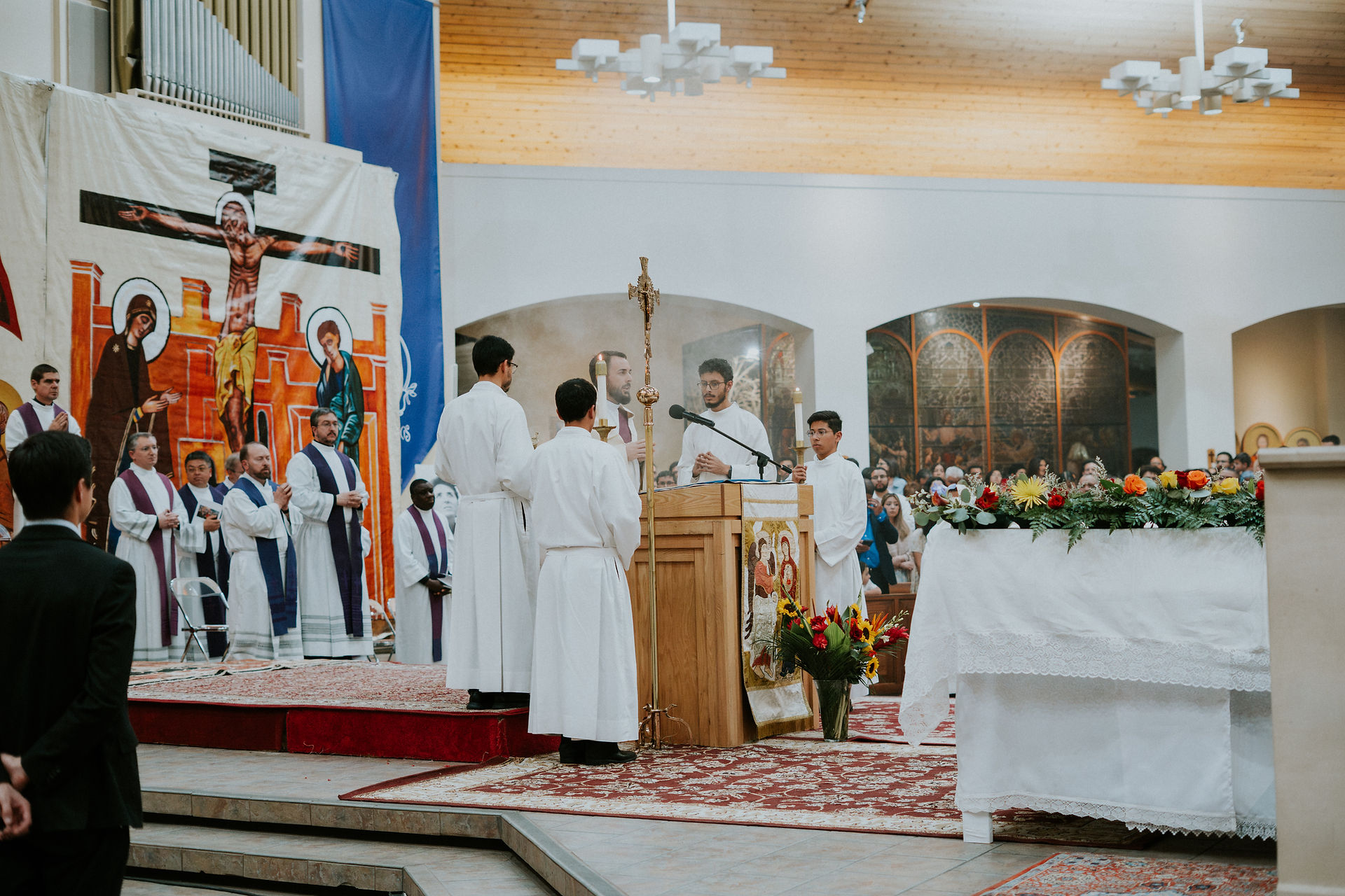Priests and altar servers stand near a decorated altar in a church service. A large religious tapestry hangs in the background.