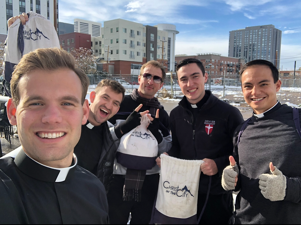 Five men in clerical attire smile outdoors, holding "Christ in the City" bags. Urban background with buildings and a clear sky.