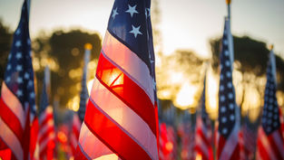 Close-up of a U.S. flag waving at sunset, with many American flags blurred in the background on a memorial-like field