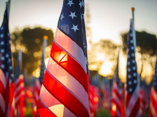 Close-up of a U.S. flag waving at sunset, with many American flags blurred in the background on a memorial-like field