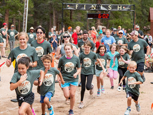 People in green shirts run in a race towards a finish line marked "FINISH" with a timer showing 6:02; trees in the background.