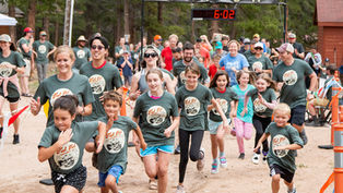 People in green shirts run in a race towards a finish line marked "FINISH" with a timer showing 6:02; trees in the background.