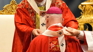 Two Catholic clerics in ornate red vestments during a solemn ceremony, one placing a stole on the other before a gold altar.