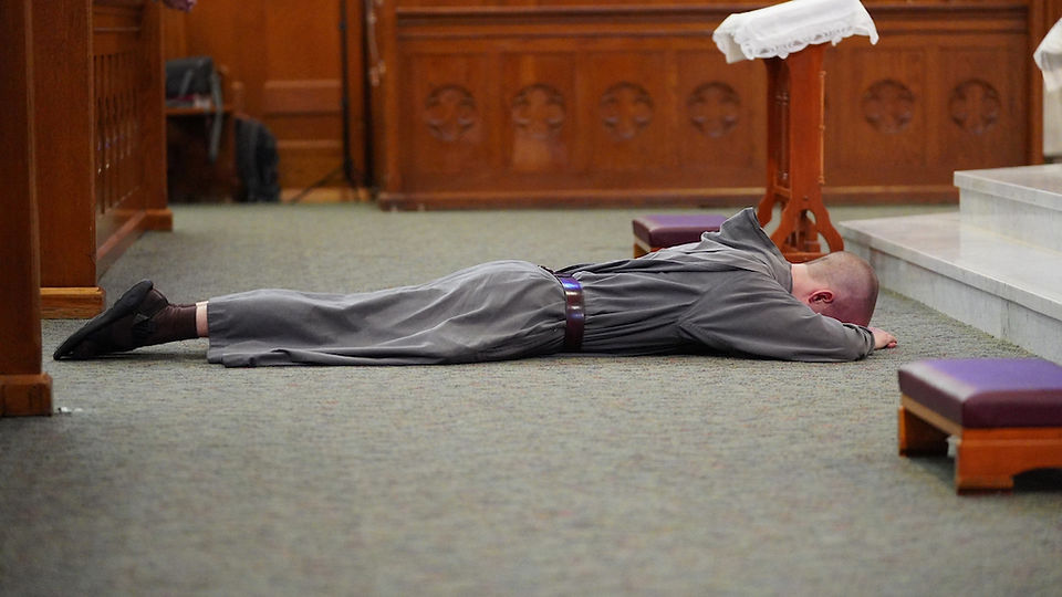 A person in a gray robe lies prostrate on a church floor near a wooden altar. The setting conveys a solemn or prayerful mood.