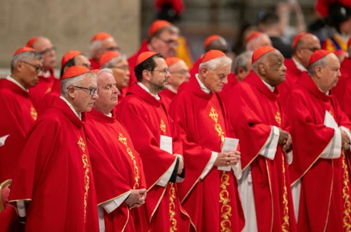 A group of cardinals in red robes stand solemnly in a church setting, holding papers. The scene is formal and reverent.