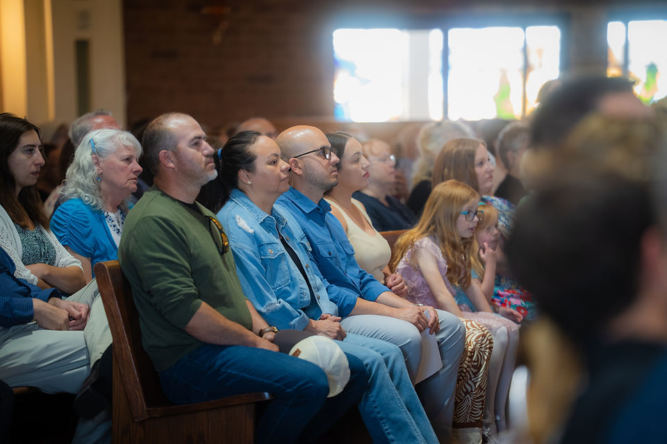 Seated church congregation of adults and children listening attentively in a bright, sunlit sanctuary.