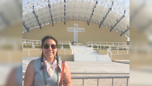 Smiling woman with sunglasses stands in front of an outdoor stage featuring a large cross; clear sky, metal beams, and white steps visible.