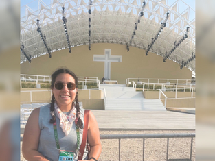 Smiling woman with sunglasses stands in front of an outdoor stage featuring a large cross; clear sky, metal beams, and white steps visible.
