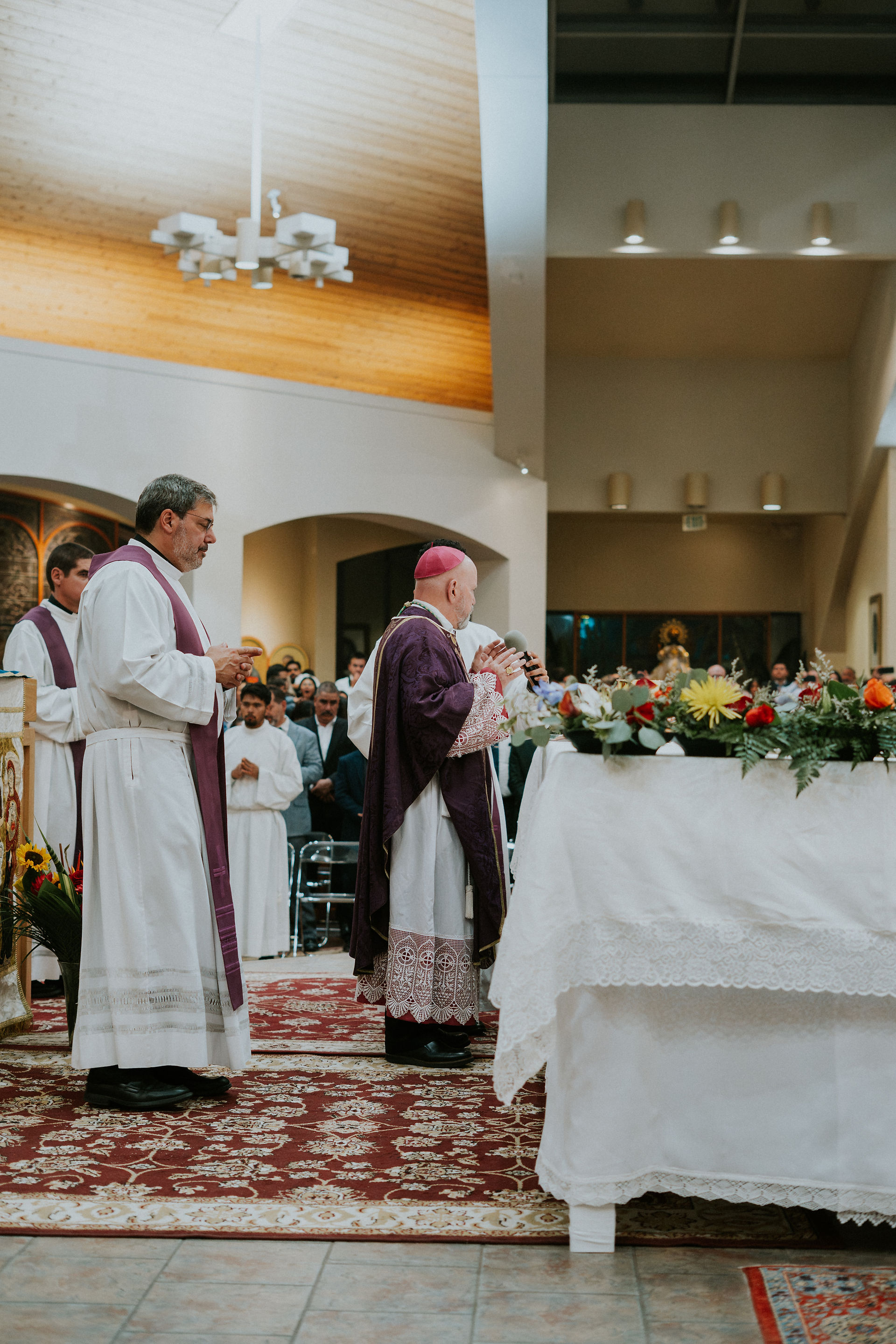 Clergy in white and purple robes stand at an altar adorned with flowers during a church ceremony. Congregation in the background.