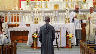 A monk in a gray robe stands before an altar in a church, with priests nearby carrying a scroll. The altar is adorned with candles and flowers.